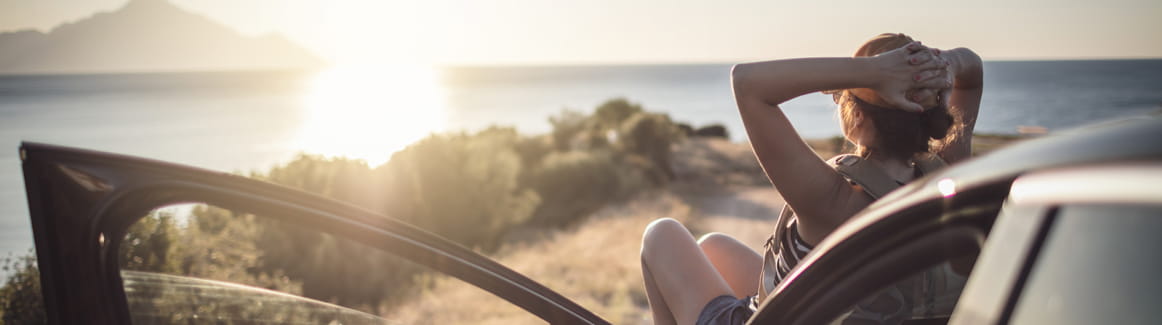 Woman sitting on the hood of her car parked at a beach watching the sunset