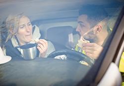 Two people in the front seat of a car, the driver eating and the passenger drinking coffee