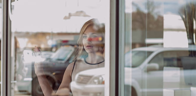 Woman standing in a window with the glare of a parking lot on the glass. 